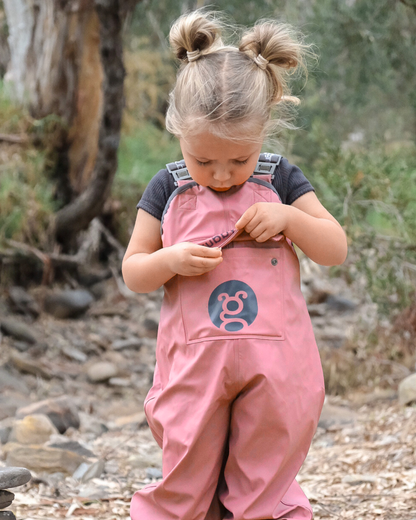 Child wearing waterproof pink overalls with a logo, standing outdoors in a natural setting.