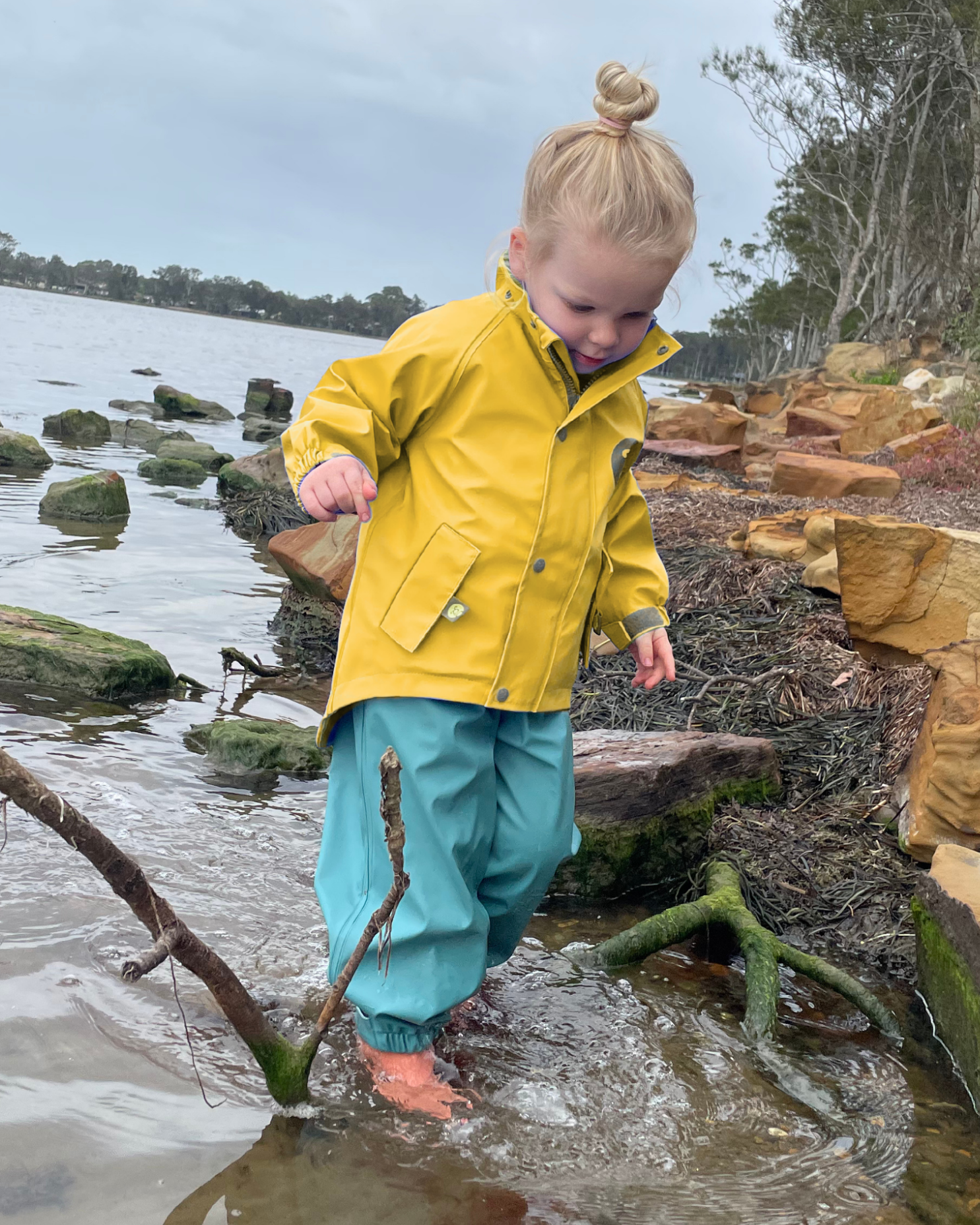 Toddler wearing yellow waterproof jacket with extra long mud guard hem walking in water at lake shoreline, kids outdoor rainwear