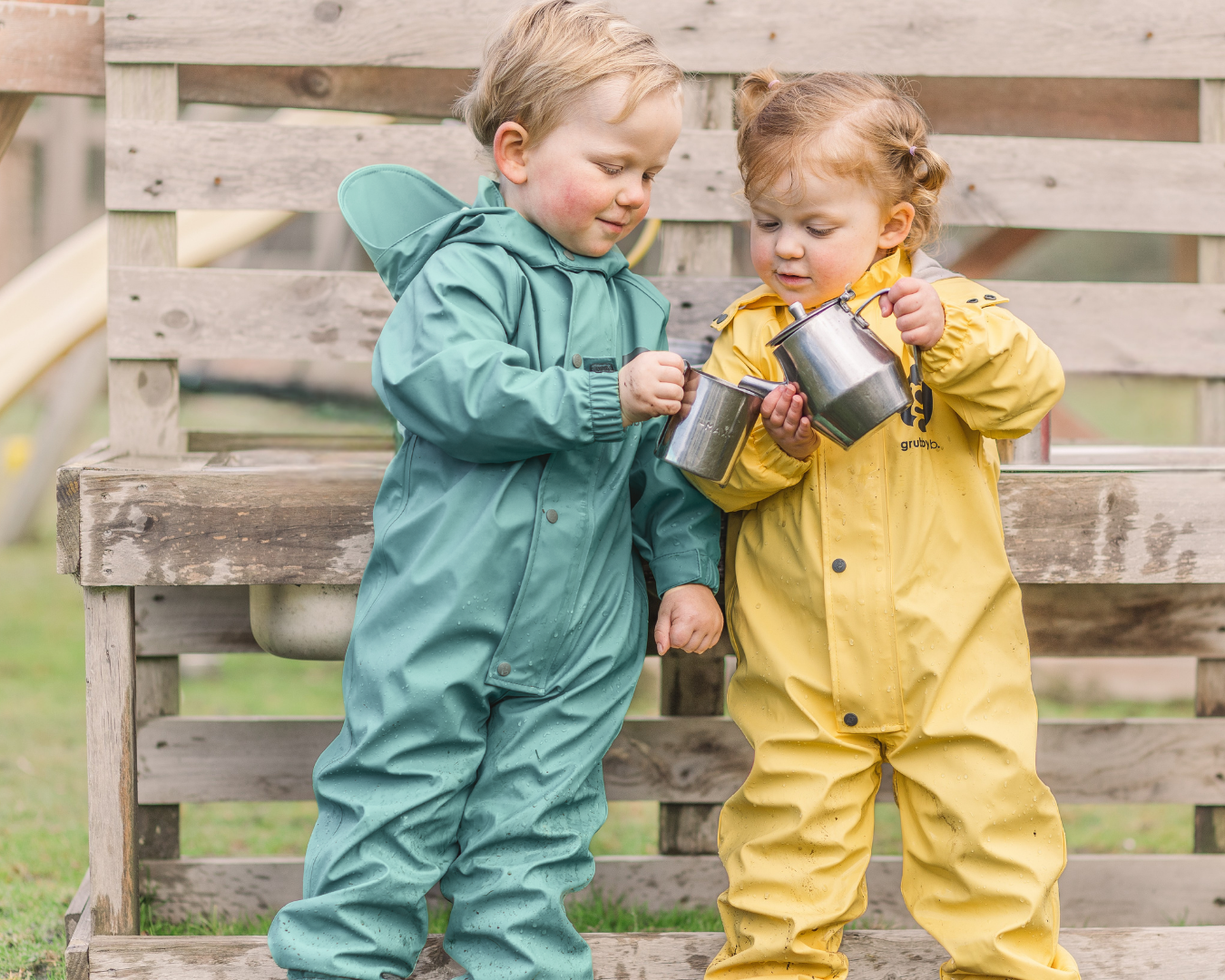 Two toddlers in waterproof rain suits playing with cups in a wooden mud kitchen, messy sensory outdoor play kids waterproof clothing