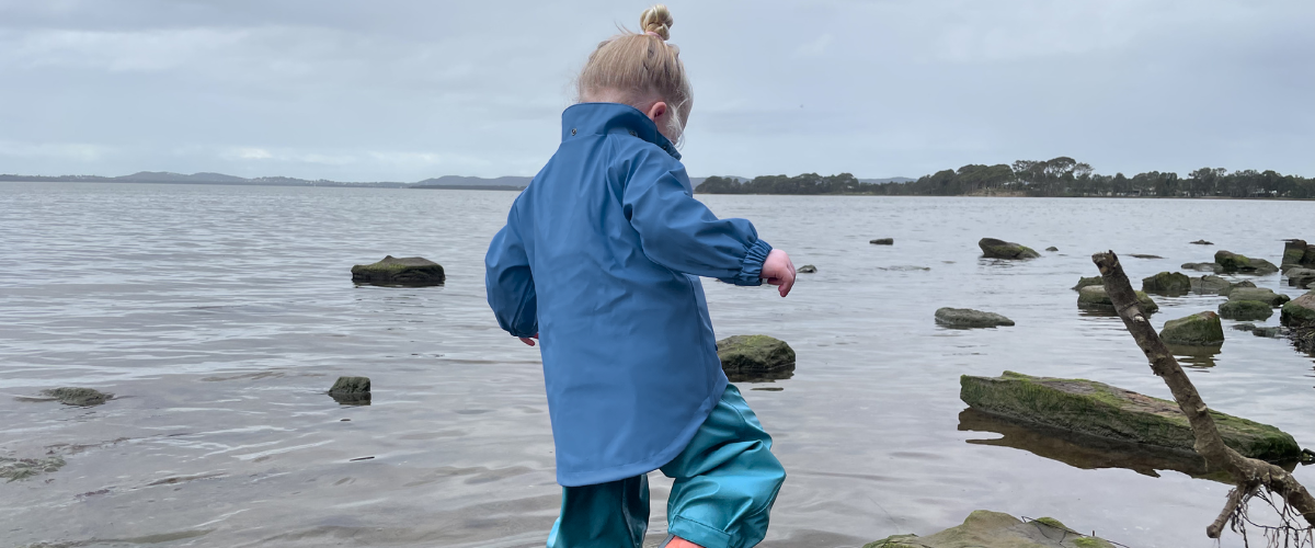 Child in a blue sustainable raincoat with extra long waterproof hem ,walking on rocks by a body of water