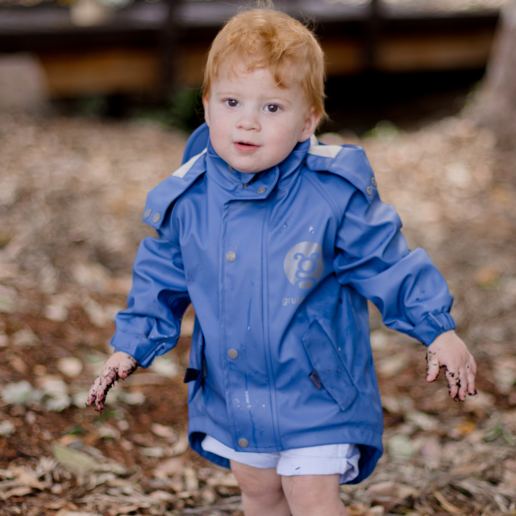 Child wearing a sustainable blue raincoat with extra long waterproof hem standing outdoors on a leafy ground.